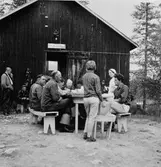 Ett större sällskap, som äter en måltid, utanför en lada.