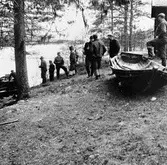 En grupp män, med eka, intill sjöstranden.