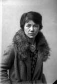 Ateljébild på en kvinna i kappa och halsband. Enligt Walter Olsons journal är bilden beställd av Ingeborg Cray ifrån USA. Kalmar läns museum känner inte till om det är den personen som är avporträtterad.