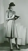 Modell i klänning med skärp, klädda knappar och fickor på bröstet, samt hatt, handskar i handen och pumps. Vit rekvisita i bakgrunden.