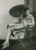 Modell i randig baddrock, sjalett, solparaply och cigarett i handen, sitter i en stol.