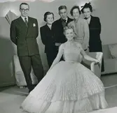 Friherrinnan Susanne Palmstierna poserar i den Diorklänning hon vunnit vid utlottning på NK:s Franska damskrädderi. Den ärmlösa klänningen har plisserat liv och kjol, midjan markerad med skärp. Underkjol av plisserad tyll. Tre kvinnor i dräkt och två män i kostym, den ene är Kurt Jacobsson i bakgrunden. NK:s Franska damskrädderi.