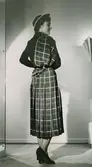 Modell i rutig klänning med veckad kjol, hatt och sjal över axeln.