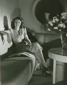 Modell i ljus klänning med draperade detaljer, halsband och ringar och högklackade skor med slingback och öppen tå, sitter i en soffa och bläddrar i en bok med en cigarett i handen.