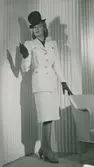 Modell i dräkt, hatt med hög kulle, handskar och pumps. Vår.