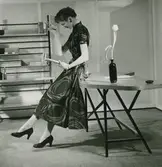 Modell i mönstrad klänning, örhänge, armband och pumps, läser i en bok, lutad mot ett bord. 