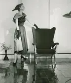 Modell i klänning med sjal och hatt, handskar och pumps, vid en fåtölj. 