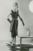 Mönster NK-Stil. Modell i klänning med skärp, pärlhalsband, handskar och pumps, vid en blomvas på ett bord. Kjolen - dräktens kjol (mönster B) Livet - aftonklänningens (mönster D)