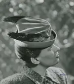 Modell i hatt med mjukt draperad kulle, uppvikt brätte. Fjädrar på sidorna.