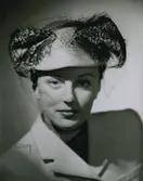 Porträtt av modell i turkosfärgad filthatt med mjuka fjädrar och flor. Av Gilbert Orcel.