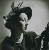 Porträtt av modell i stråhatt med fågeldekoration och flor, pärlhalsband.