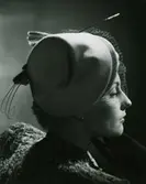 Porträtt av modell i beige filthatt med ett skärmliknande underbrätte, fantasi i brun fjäder. Av Simone Cange.