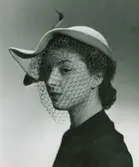 Porträtt av modell i naturfärgad filthatt med brun rosett och flor. Av Jacques Fath.
