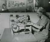 Kvinna i arbetsrock vid en industrisymaskin av märket Reece. Text med blyerts på baksidan: 