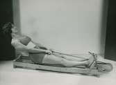Kvinna i tvådelad baddräkt tränar i en roddapparat. Nordiska Kompaniet. Text med blyerts på baksidan: 