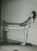 Demonstration av en massageapparat. Nordiska Kompaniet. Kvinna i hel baddräkt med massagebandet runt stussen. Text med blyerts på baksidan: 