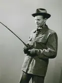 Man iklädd hatt, rutig halsduk samt en skinnjacka från C O Ericson, Malung. I handen håller han ett fiskespö. Nordiska Kompaniet. Text med blyerts på baksidan: 