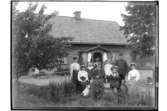 Envånings bostadshus med förstebro med snickarglädje.
Åtta personer och två hundar framför huset.
Aug. Karlsson