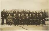 Fotografiskt vykort av grupp män, varav de flesta är militärer, andra civila. Stämpeln på kortets baksida (