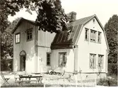Bostadshus med frontespis och mansardtak, året är 1908. På tomten finns trädgårdsmöbler.