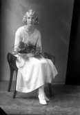 Ateljébild på en kvinna i klänning ihållandes en blombukett. Enligt Walter Olsons journal är bilden beställd av Stina Jansson.