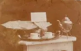 Vykort skrivet från Ellen Warholm till Karin Warholm 1917, från Borlänge till Falun. Bilden Föreställer ett kaffebord med kaffepetter, två koppar och gräddkanna, men sockerskål och kakfat är fulla av ransoneringskort. Överst ettvitt kort med texten: 