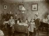 Familjen Gustav Johansson samlade kring bordet i hemmet. Till vänster mamma Maria Lovisa, född Nilsson med sonen Bengt i Knät. Till höger pappa Gustav med dottern Signe i knät. Tapeter, bordsduk och övrig inredning är jugendmönstrade.