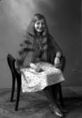 Ateljébild på en flicka i klänning sittandes på en pall. Enligt Walter Olsons journal är bilden beställd av fröken Amy Gustavsson.