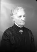 Ateljébild på en kvinna i hårhuppsättning och brosch. Enligt Walter Olsons journal är bilden beställd av frun till rektor Allmér.