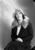 Ateljébild på en kvinna i kappa och hatt. Enligt Walter Olsons journal är bilden beställd av fru M Håkansson.