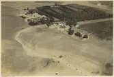 Bild högt uppifrån (flygbild?) över fält, väg och oidentifierad byggnad. Stämpeln på kopians baksida (