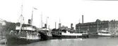 Ångbåtar (Kalmarsundsfärjor ) med vimplar i Ölandshamnen utanför margarinfabriken i Kalmar. Nuvarande Baronens köpcentrum medan hamnen är Kalmar gästhamn.
