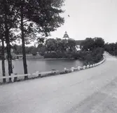 En väg med ett räcke bredvid en sjö. I bakgrunden syns kyrkan i Hjorted.