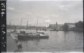 Båtar förtöjda vid Museikajen i Stockholm. I bakgrunden Skeppsholmen, Strandvägen och Djurgården med Nordiska museet. Motorjakten med ljust skrov till v (skymd) är den C. G. Pettersson-ritade TATJANA (1921); i bakgrunden till h skymtar postångaren ÖLAND (b. 1883, reg nr 3889).