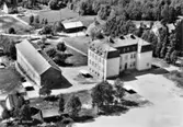 Flygbild över Ala skola med lågstadiebyggnaden till vänster och mellanstadiebyggnaden till höger. Skolvaktmästarbostaden ses nederst till vänster. Vykort.