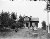 Man liggande i gräset, barn och kvinnor sitter på bänken, framför bostadshus, Östhammar, Uppland