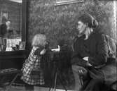 Kvinna och barn med docka.
Fotografen Nanny Ekström kan skymtas i spegeln med sin kamera.