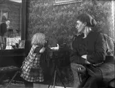 Kvinna och barn med docka.
Fotografen Nanny Ekström kan skymtas i spegeln med sin kamera.
