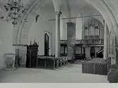 Interiör av okänd kyrka, sett från koret över bänkarna mot orgelläktaren. Till vänster i bild dopfunt, dörr till predikstol och port, till höger skymtar kaminen.
