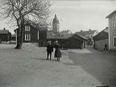 Stadsvy med Strängnäs kyrktorn i bakgrunden. I förgrunden två finklädda barn på en platsbildning med pump.