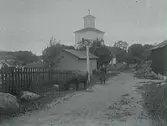 Okänd kyrka på okänd ort (samma kyrka som på neg. nr. E5516). I förgrunden en lösgående betande ko bland bebyggelsen samt en flicka vid gatlykta bakom kon.