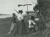 Konstnär vid staffli - ser ut att vara Severin Nilson själv - under parasoll i havsmiljö med två kvinnor som studerar hans måleri.