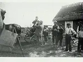 Mannen i vagnen är på besök och gårdens folk bjuder på kaffegök. Tre män, varav två dricker ur pilsnerflaskor; den ene håller en tallrik med ett glas. En av de fyra barfota gossarna håller en liten hundvalp. till vänster i bild står ett staffli och bakom dem syns stugan med halmtak.