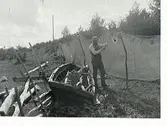Man som lagar fiskenät. Intill honom ligger en båt på land mot en hankgärdsgård.