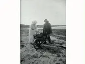 Fiskare med kärra på en vid sandstrand och en finklädd kvinna som böjer sig över vagnen, troligen en sommargäst.