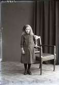 Flickporträtt, flicka stående i helfigur vid länsstol. Ateljébild. Beställare: Wera Mossberg. Fototid (osäker) 1910.
