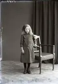 Flickporträtt, flicka stående i helfigur vid länsstol. Ateljébild. Beställare: Wera Mossberg. Fototid (osäker) 1910.