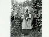 Kvinna i sockendräkt med psalmbok och blomsterbukett (liljekonvalj?). Hon står i en grindöppning.