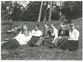 Gruppbild. Sju ungdomar sitter i en solig skogsbacke. En av ynglingarna har käpp och tjejen intill honom håller i en pinne.