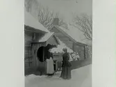 Två damer vid skunken till en högloftsstuga. Snöbild.